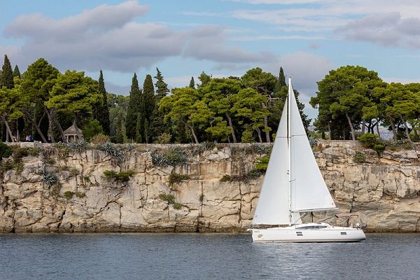 Image of a sailboat with white sails against the backdrop of cliffs overgrown with vegetation.