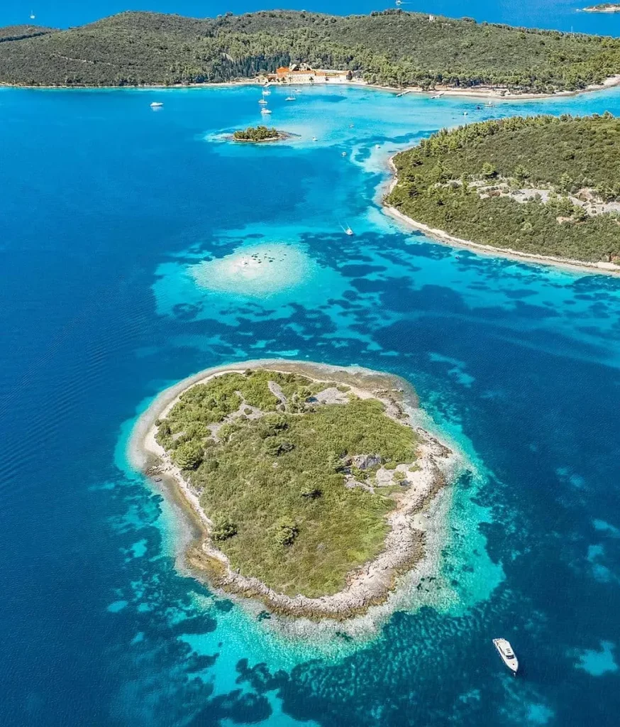 The image showcases a cluster of islands bathed in vibrant sunlight. The crystal-clear, turquoise water reveals a mosaic of sandy shallows and deeper azure depths.  A lush, green island anchors the center of the scene, its untouched vegetation creating a sense of tranquility. Several other islands, varying in size, dot the seascape, their shores fringed with trees.

Numerous sailboats and yachts are anchored or gently gliding across the calm waters, their white hulls contrasting with the vivid blue sea. A luxurious white yacht, its sleek design catching the sunlight, sits prominently near the central island.