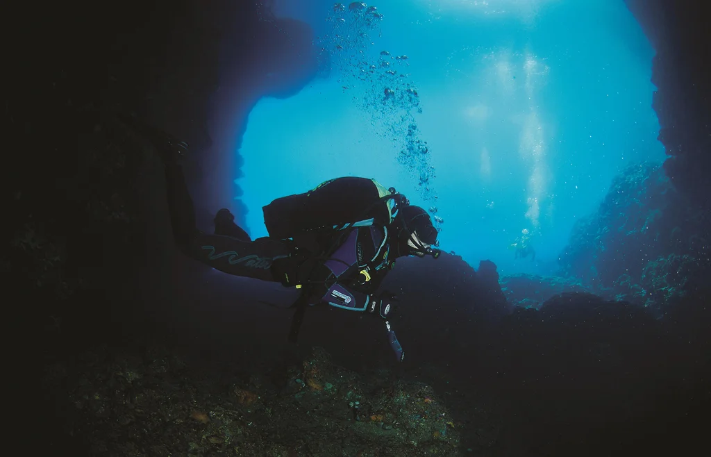 An underwater image of a diver in full diving gear. It's taken in an underwater cave - the diver is looking at the sea bed with a flash light. 