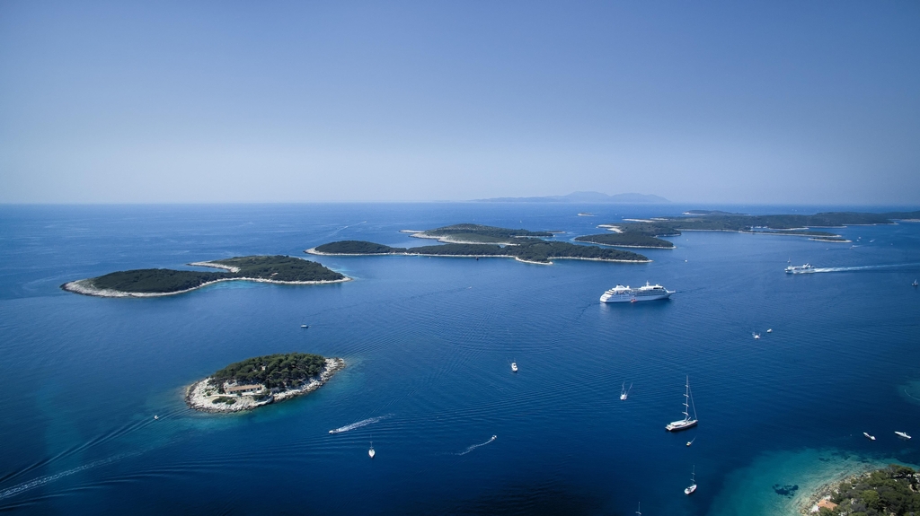 The image showcases a stunning vista of a vibrant blue Adriatic Sea, its surface shimmering under a bright sun. Multiple islands, varying in size and shape, dot the seascape, their lush green vegetation contrasting beautifully with the azure waters. Some islands boast rocky coastlines, while others have inviting sandy beaches.

Several boats, ranging from small sailboats to luxurious yachts, navigate the calm waters, leaving gentle wakes in their paths. A large cruise ship, a symbol of leisure and travel, anchors majestically in the deeper waters, adding a sense of scale to the scene.