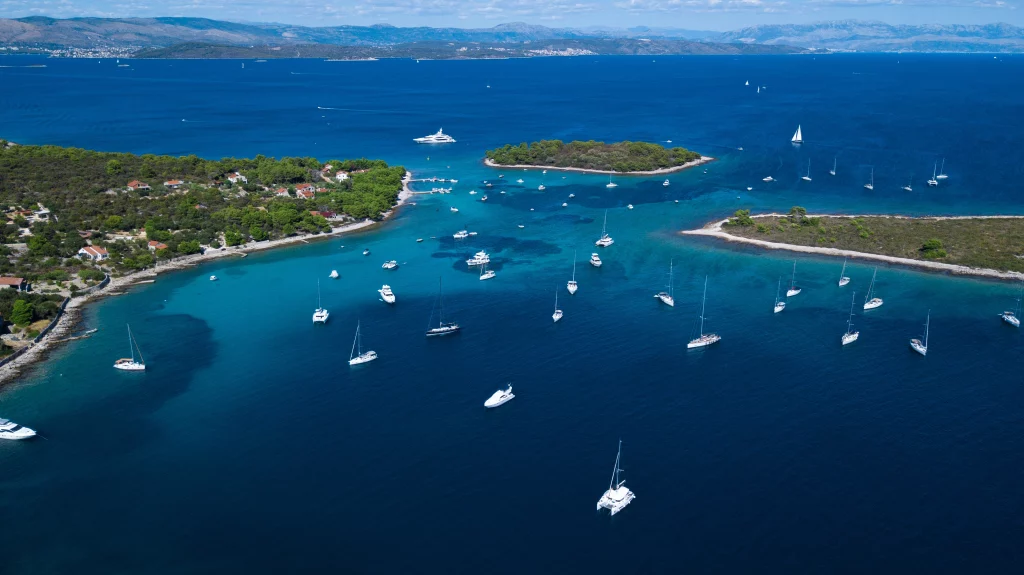 An aerial photograph showcasing the stunning Blue Lagoon in Croatia. The vibrant turquoise water contrasts with the lush green vegetation on the surrounding land. Several boats are anchored in the calm waters, and a small, idyllic island can be seen in the background under a bright blue sky with fluffy white clouds.