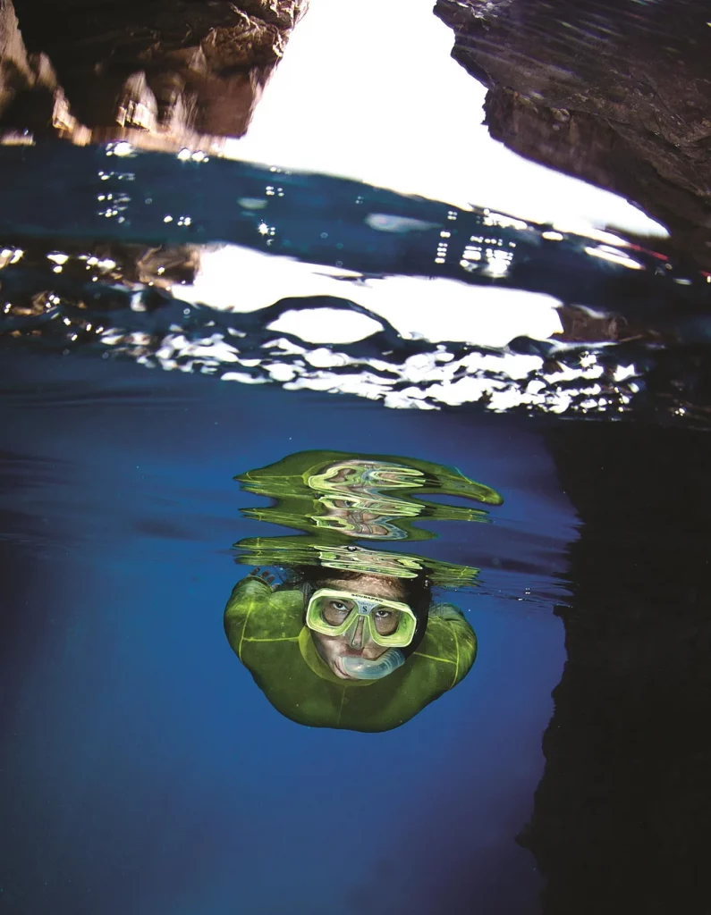 Image of a woman snorkeling in deep water. The woman is wearing a green dive suite and a green mask and a snorkeler. The shot is taken under water with the woman breaking the water and gliding through it while looking straight into the camera.