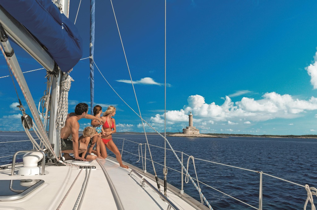 A group of people with children on a deck of a sailboat on a clear summer day. In the forefront is the bow of the ship and the sea with small waves, and in the distance is flat land with a stone lighthouse jutting up.