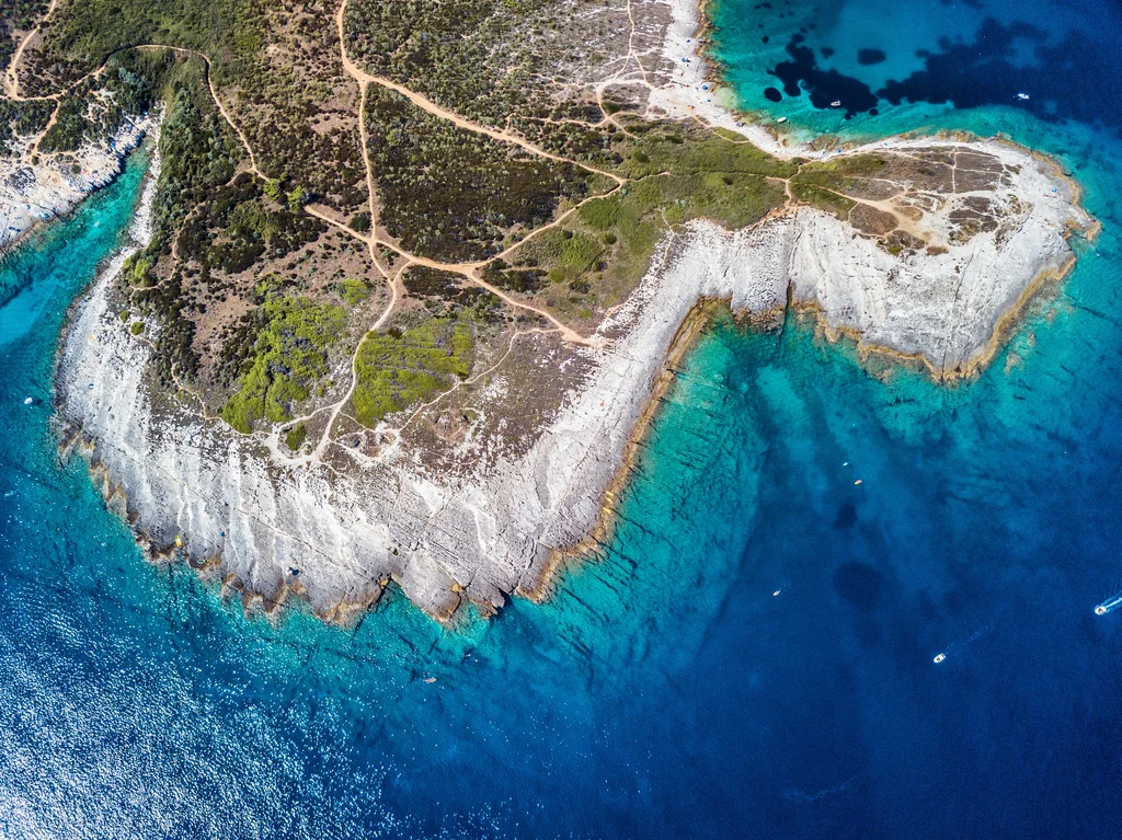 Aerial photo of Cape Kamenjak Istria. Top of the image features a landmass with various shades of green and brown and white cliffs descending into the sea. The bottom of the photo features crystal clear Adriatic Sea - high visibility allows a view of the seabed. A few motor boats can be on the surface of the sea.