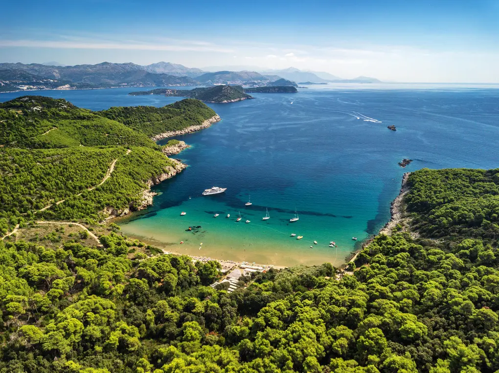 Aerial perspective of a picturesque coastal scene. Lush green forest blankets the land, opening up to a crescent-shaped beach with pale golden sand. Calm, turquoise water fills the bay, dotted with white sailboats and motorboats. The horizon stretches out to reveal distant islands and hazy mountains under a bright blue sky. The scene evokes feelings of tranquility, warmth, and the gentle sounds of the sea.