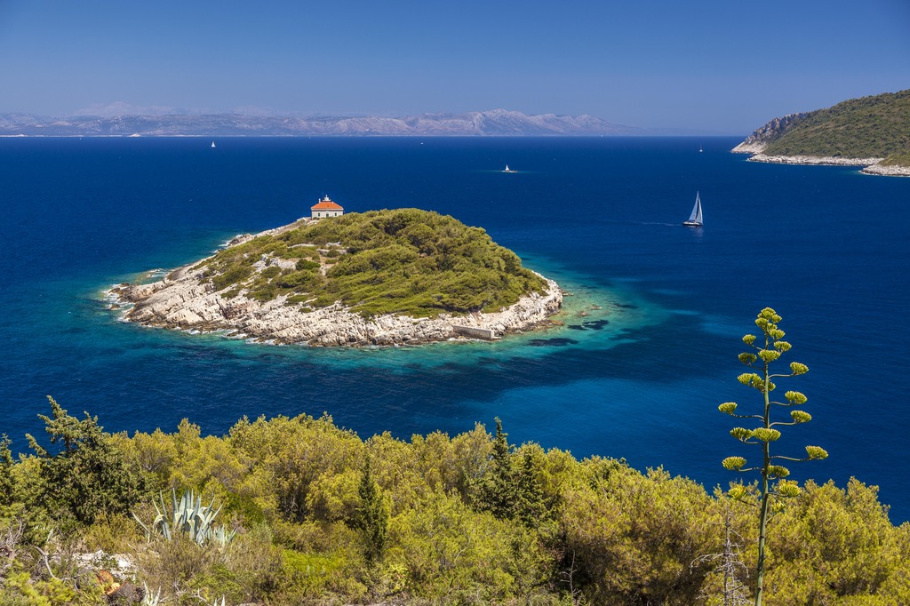 A small island surrounded by a deeply blue sea dotted with sailboats and tall mountains on the horizon.