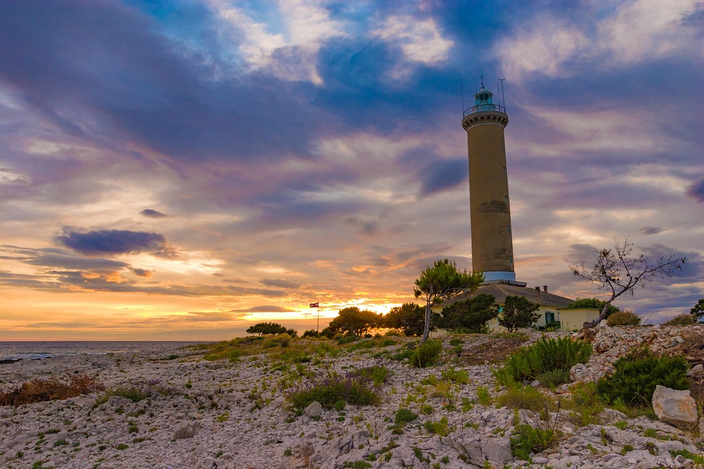 Lighthouse shown in the distance against a backdrop of a cloudy sky during sunset.